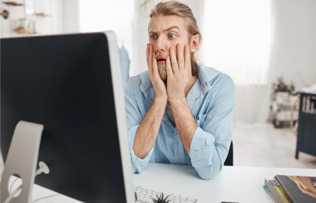 Dissatisfied young male manager looking with bugged eyes and astonishment, shocked by financial report, leaning on elbows while sitting at table in front of computer screen during hard working day